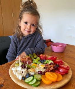 Child wearing a navy blue sweater sitting at a table with a plate of food in front of him. There are tomatoes, cucumbers, olives, grapes, cheese, bacon, and eggs.